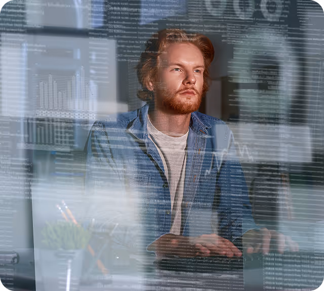 Young man with red hair and beard, wearing a denim jacket, working on a computer with transparent data and graphs overlay.