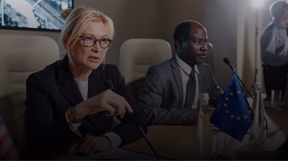 Two professionals seated at a conference table with a European Union flag, speaking into microphones.