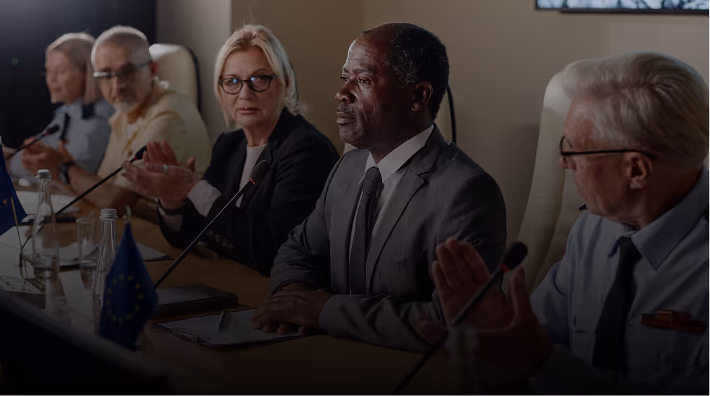 A diverse group of professionals sitting around a conference table, with some clapping, during a formal meeting.