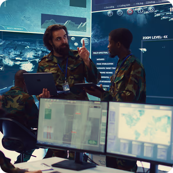 Two military personnel in camouflage uniforms discussing with tablets in a command center with digital maps and data screens.