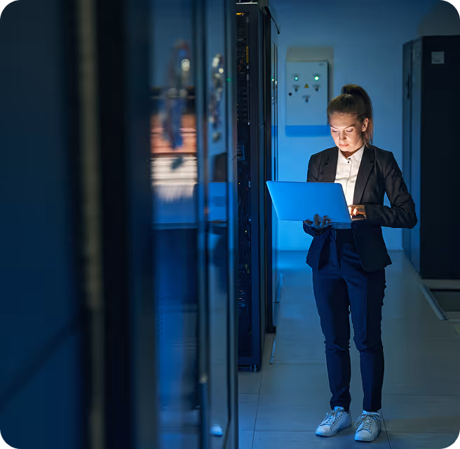 Woman in business attire working on a laptop in a dimly lit server room.