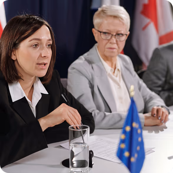 Two professional women seated at a meeting table with documents and a European Union flag in front of them.