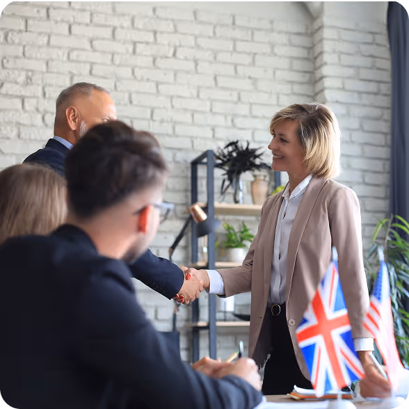 Businesswoman smiling and shaking hands with a man in an office setting with British and American flags on a table.