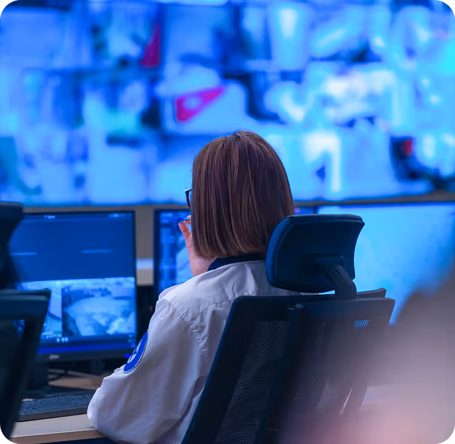 Person with short hair sitting in an office chair facing multiple monitors displaying security footage.