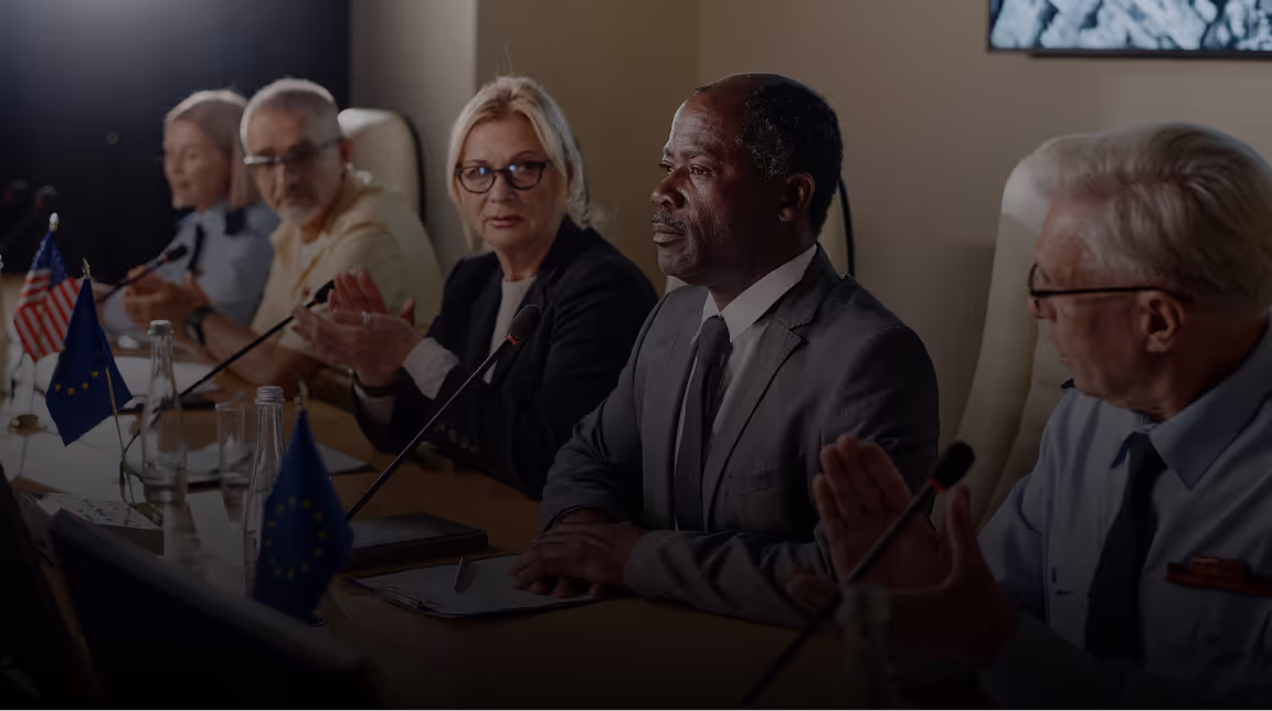 Diverse group of professionals seated at a conference table with microphones and flags, engaged in a discussion.