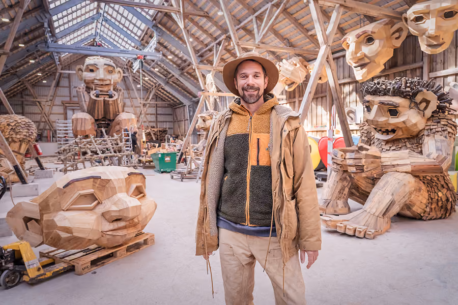 Man wearing a hat and outdoor jacket standing inside a wooden workshop surrounded by large, intricate wooden sculptures of human-like faces and figures.