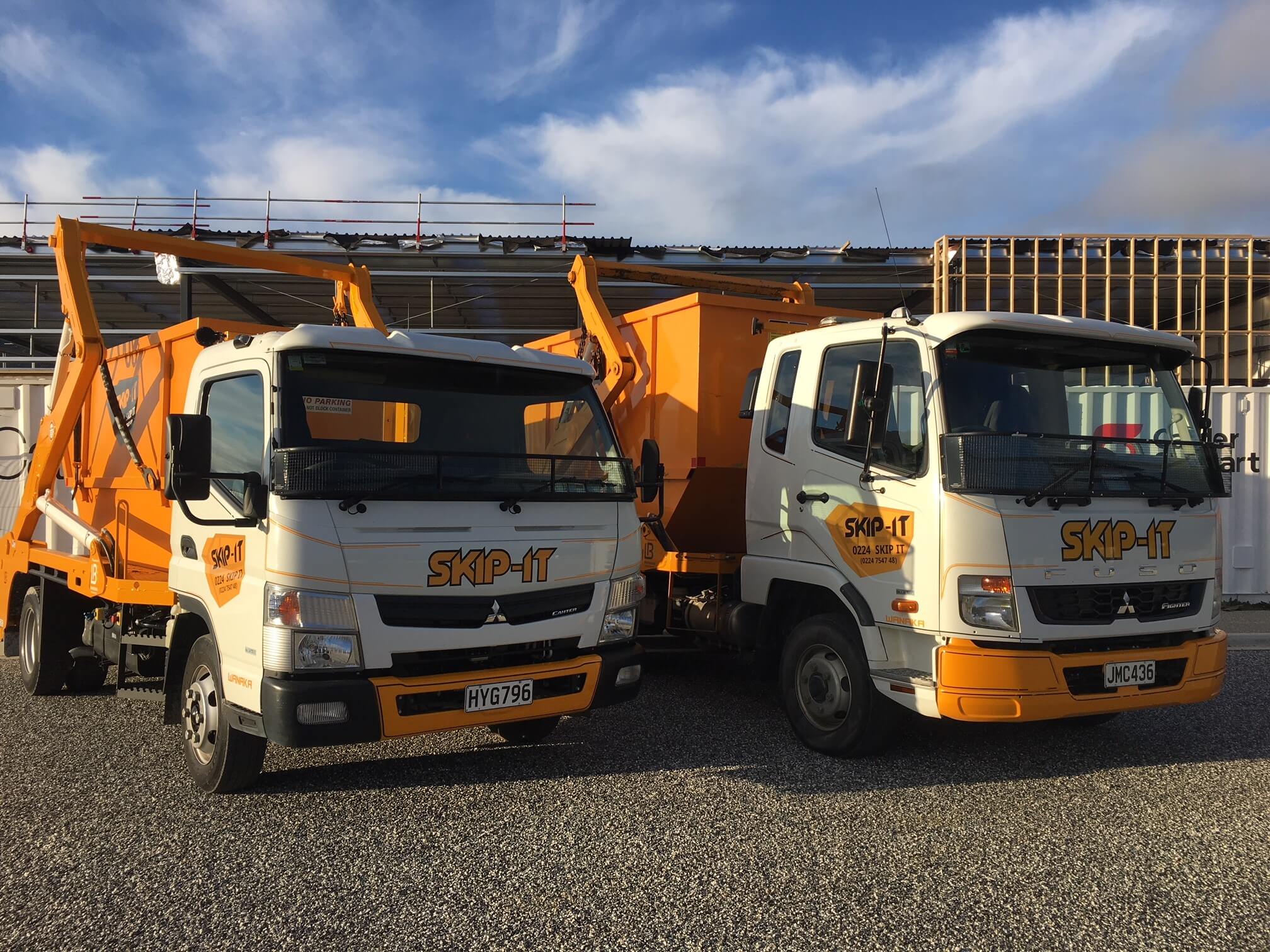 Two white and orange skip bin trucks parked on gravel in front of a construction site with a partly cloudy sky overhead; both trucks have the logo 'SKIP-IT' on their fronts and sides.