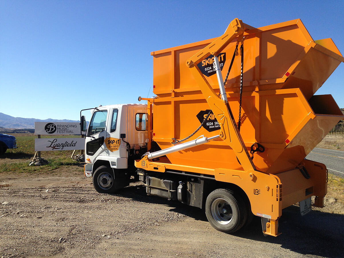White skip truck carrying two stacked bright orange skip bins on a dirt area under a clear blue sky near a road with signs for Branding Music Festival and Lyonfield.