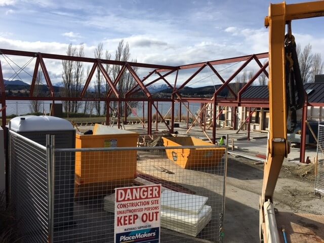 Construction site with steel framework in progress near a body of water and mountains in the background, fenced with a sign reading 'Danger Construction Site Unauthorized Persons Keep Out'.