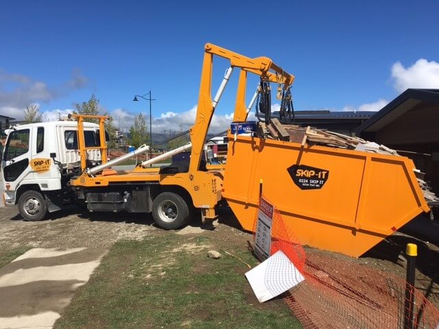 Orange and white skip bin truck unloading a large orange skip bin filled with wooden debris next to a construction site under a clear blue sky.