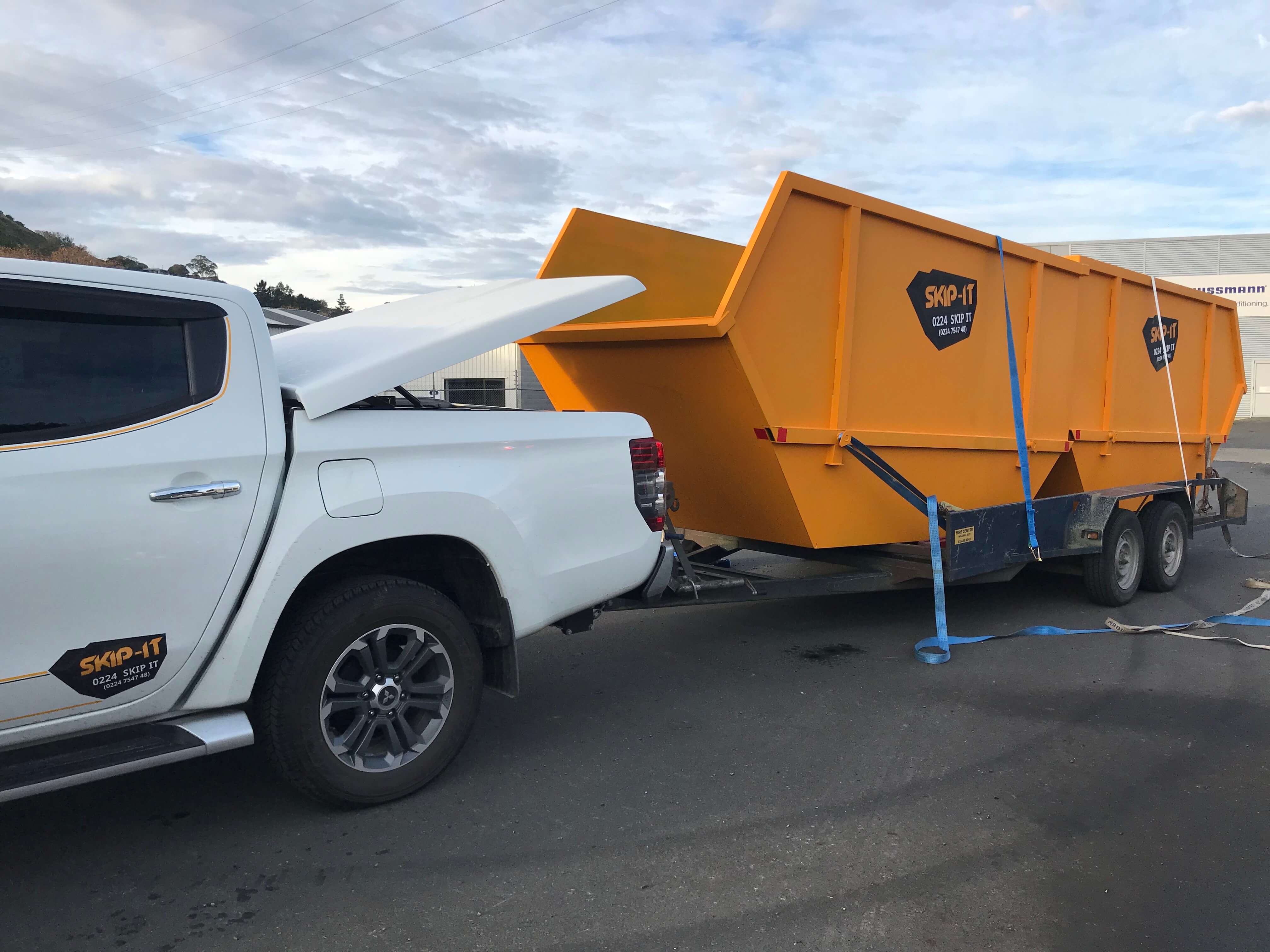 White pickup truck with its rear tonneau cover open, towing a large yellow skip bin secured on a dual-axle trailer, both branded with 'SKIP-IT' logos, parked on asphalt with a cloudy sky background.