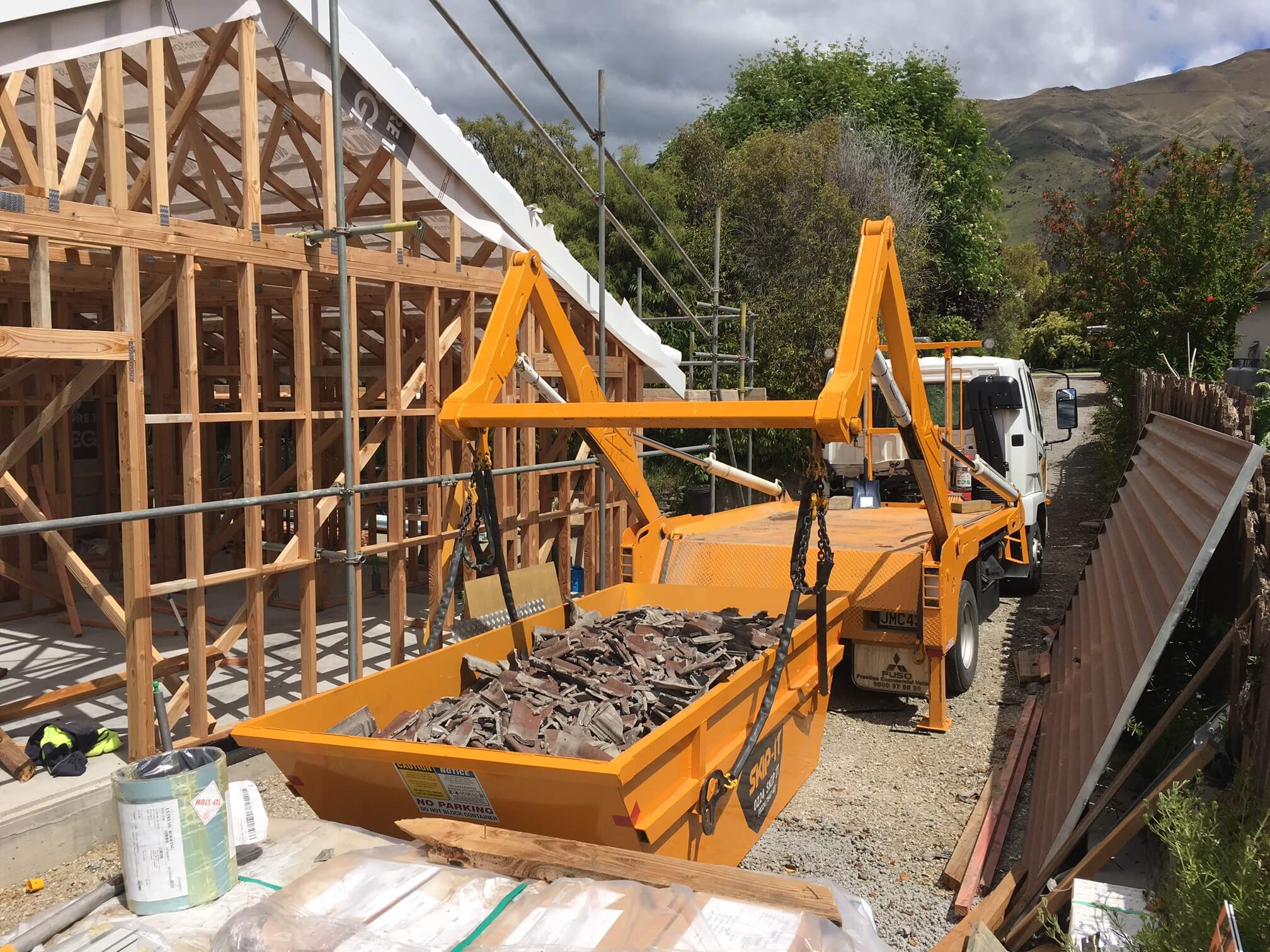 Construction site with a wooden frame of a house under a cloudy sky and a white truck carrying a bright orange skip container filled with broken roof tiles.