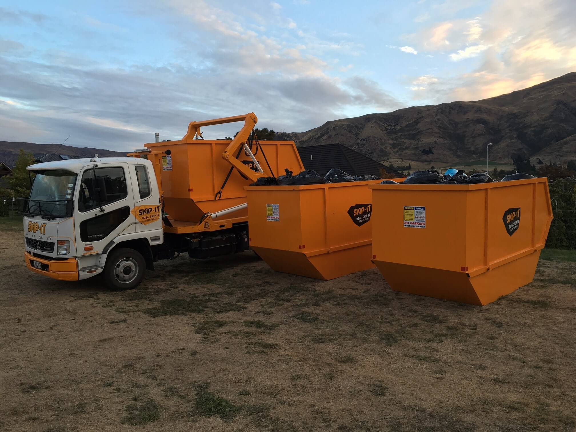 A white and orange skip truck labeled 'SKIP-IT' parked on dry ground, with two orange skip bins full of black garbage bags beside it, set against a backdrop of mountains and a cloudy sky at sunset.