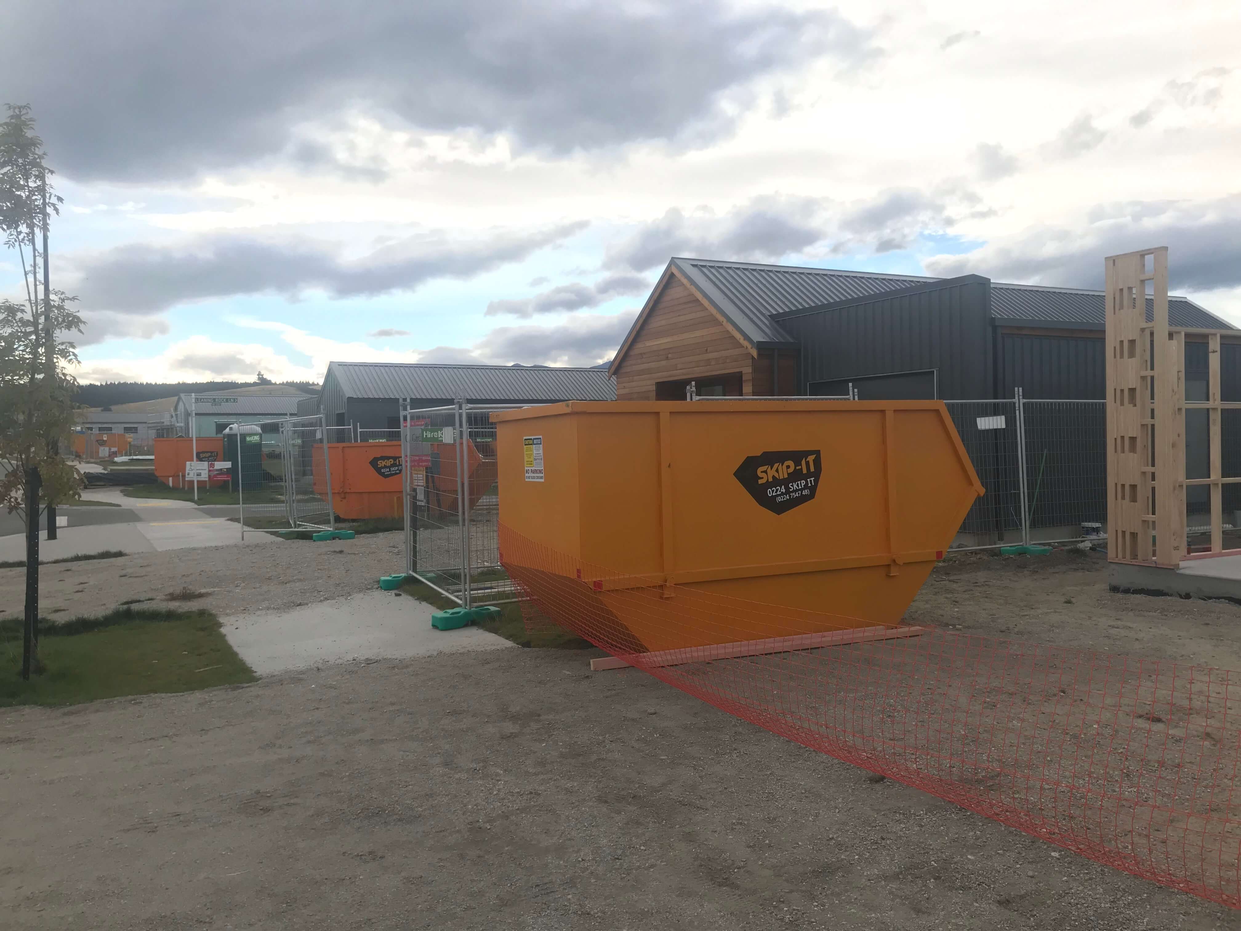 Construction site with several orange skip bins labeled 'SKIP-IT' lined up on gravel beside buildings with metal roofs under a cloudy sky.