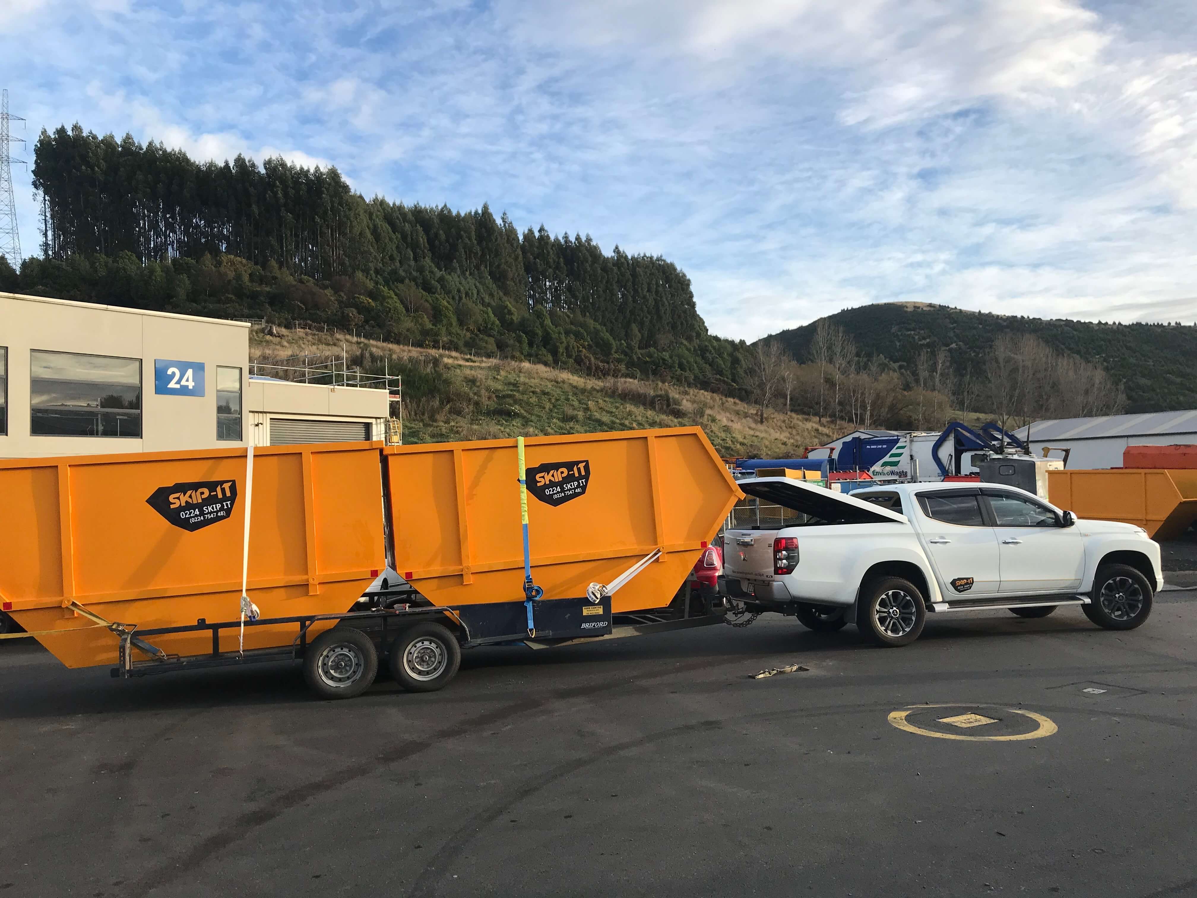 White pickup truck towing a double-axle trailer with two large orange skip bins labeled 'SKIP-IT' parked in an industrial area with buildings, trees, and green hills in the background.