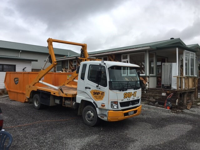 A white and orange Skip-It branded Fuso truck with a large skip container is parked in front of a house undergoing demolition or renovation, with part of its exterior wall removed.