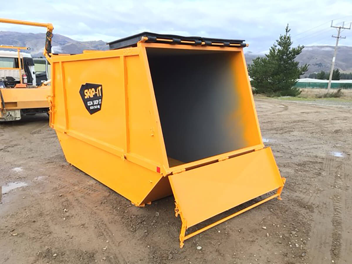 Large yellow metal skip bin with an open front door, placed on a dirt surface with mountains and trees in the background.