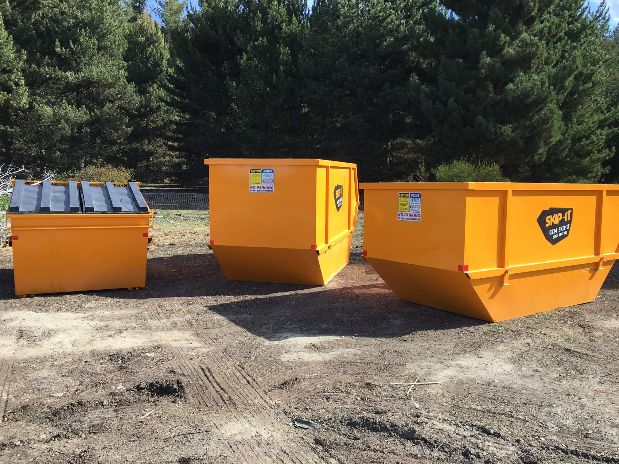 Three bright yellow skip bins of different sizes placed on bare ground with a backdrop of green pine trees under a clear sky.