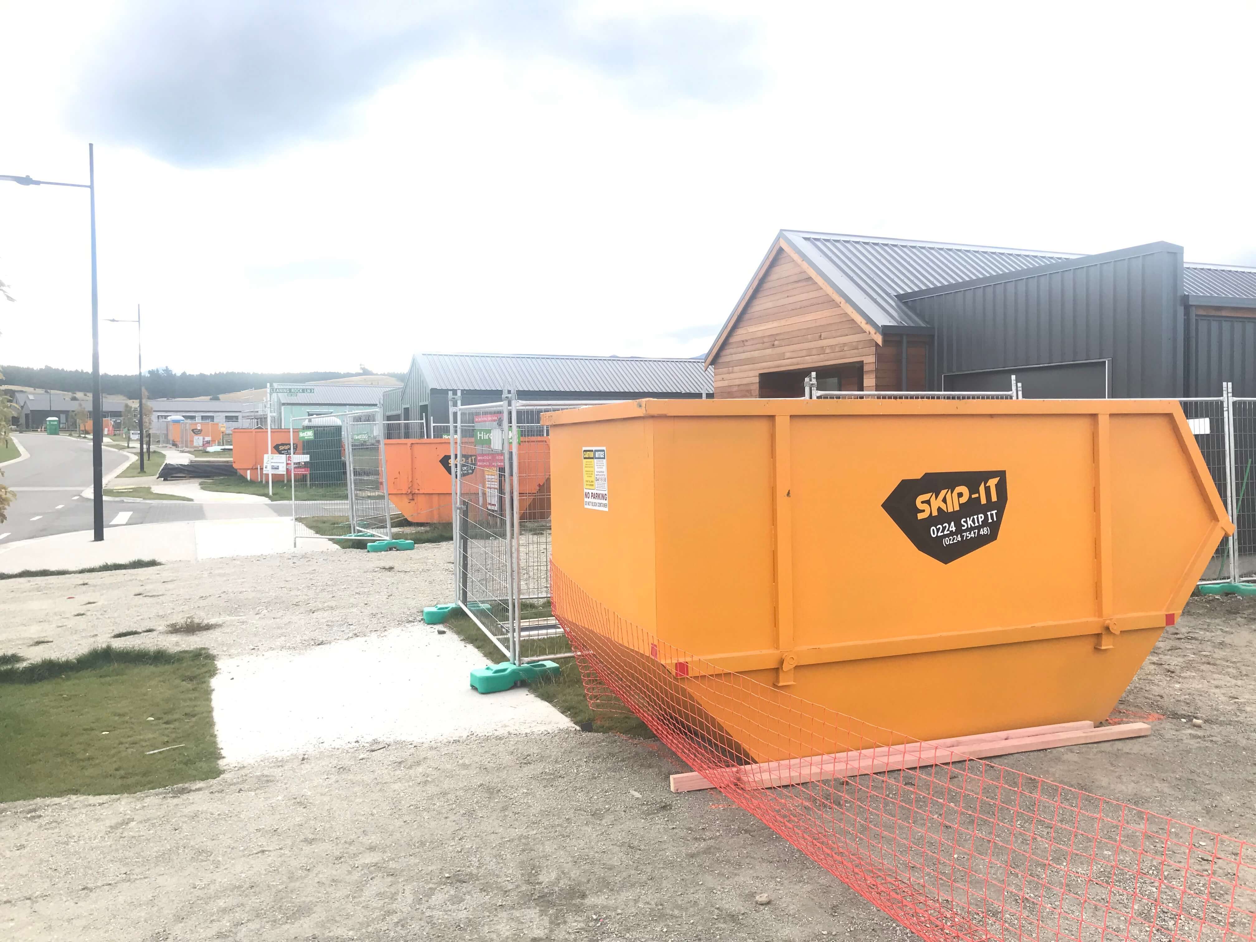 Multiple large orange skip bins branded 'SKIP-IT' lined up along a fenced construction site sidewalk near modern industrial-style buildings.