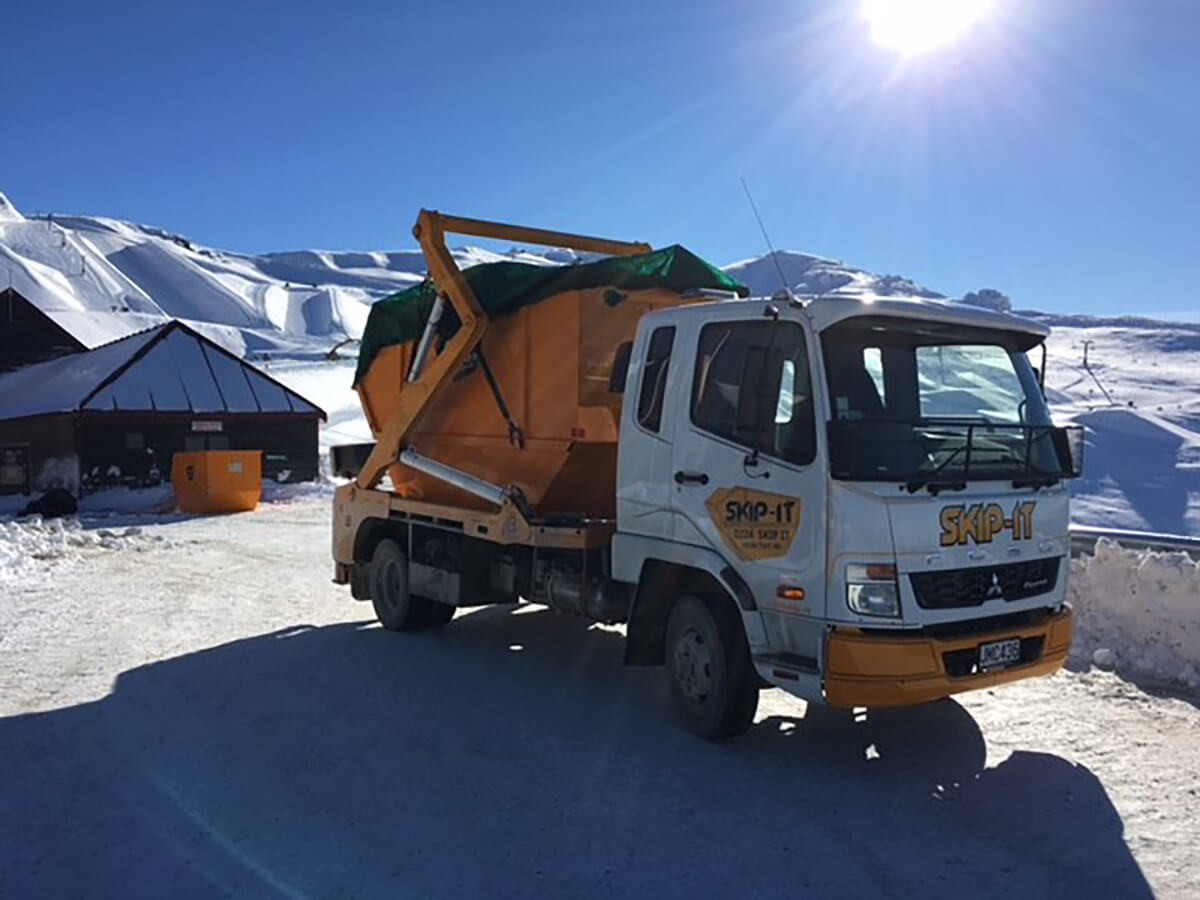 White and orange Skip-It truck parked on a snowy road with snow-covered buildings and mountains in the background under a clear, sunny sky.