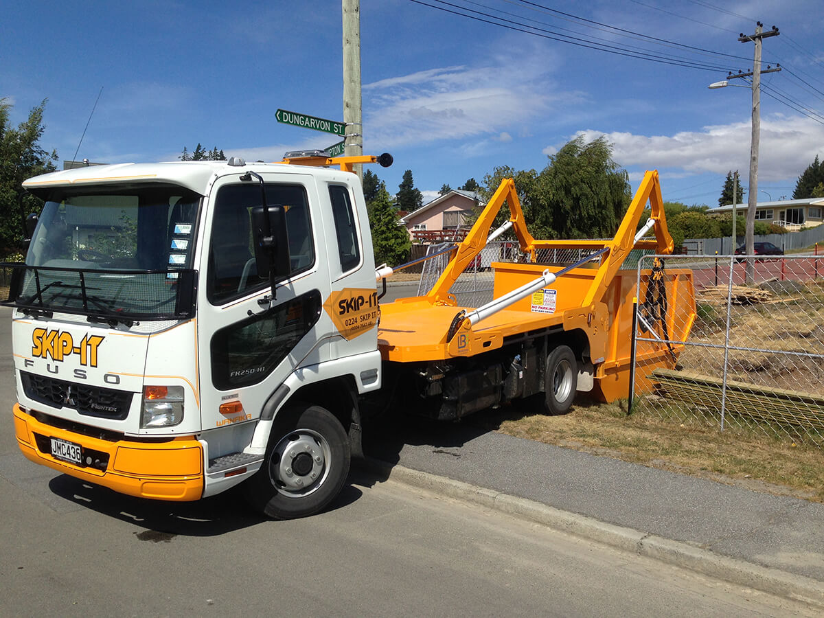 White and orange Skip-It Fuso flatbed truck parked on a street next to a fence and grassy area with street signs visible in the background under a partly cloudy blue sky.