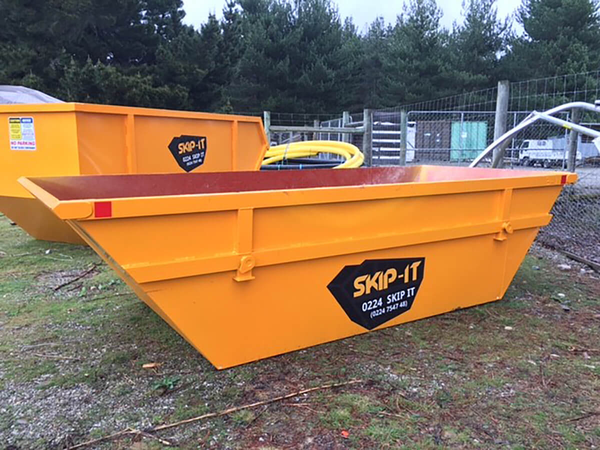 Two large yellow metal skip bins labeled 'SKIP-IT' placed outdoors on grass with trees and a fenced area in the background.