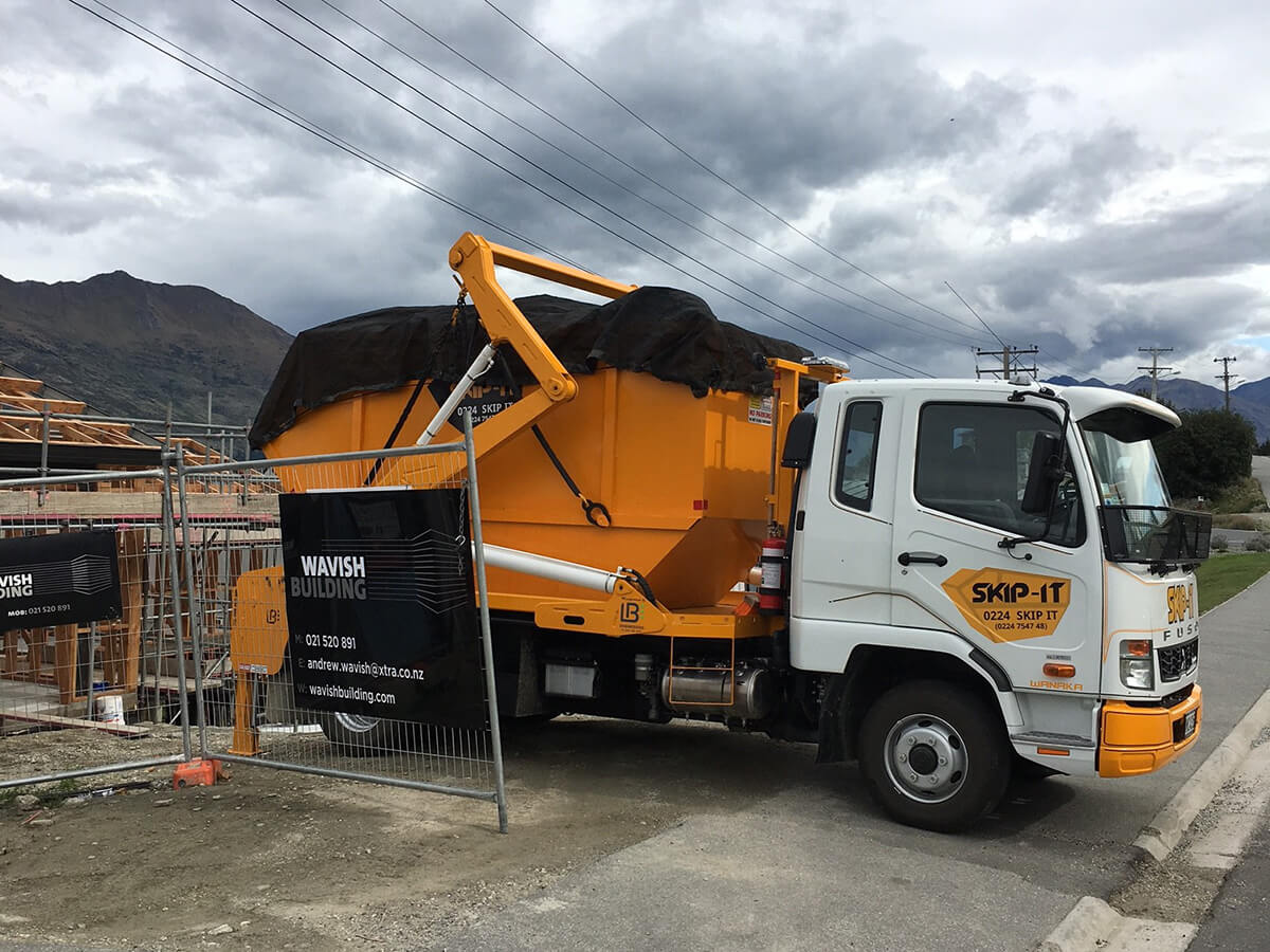 White and yellow skip bin truck labeled SKIP-IT parked by a construction site with a black-covered skip bin on its back, fenced by Wavish Building banners under cloudy sky with mountains in background.