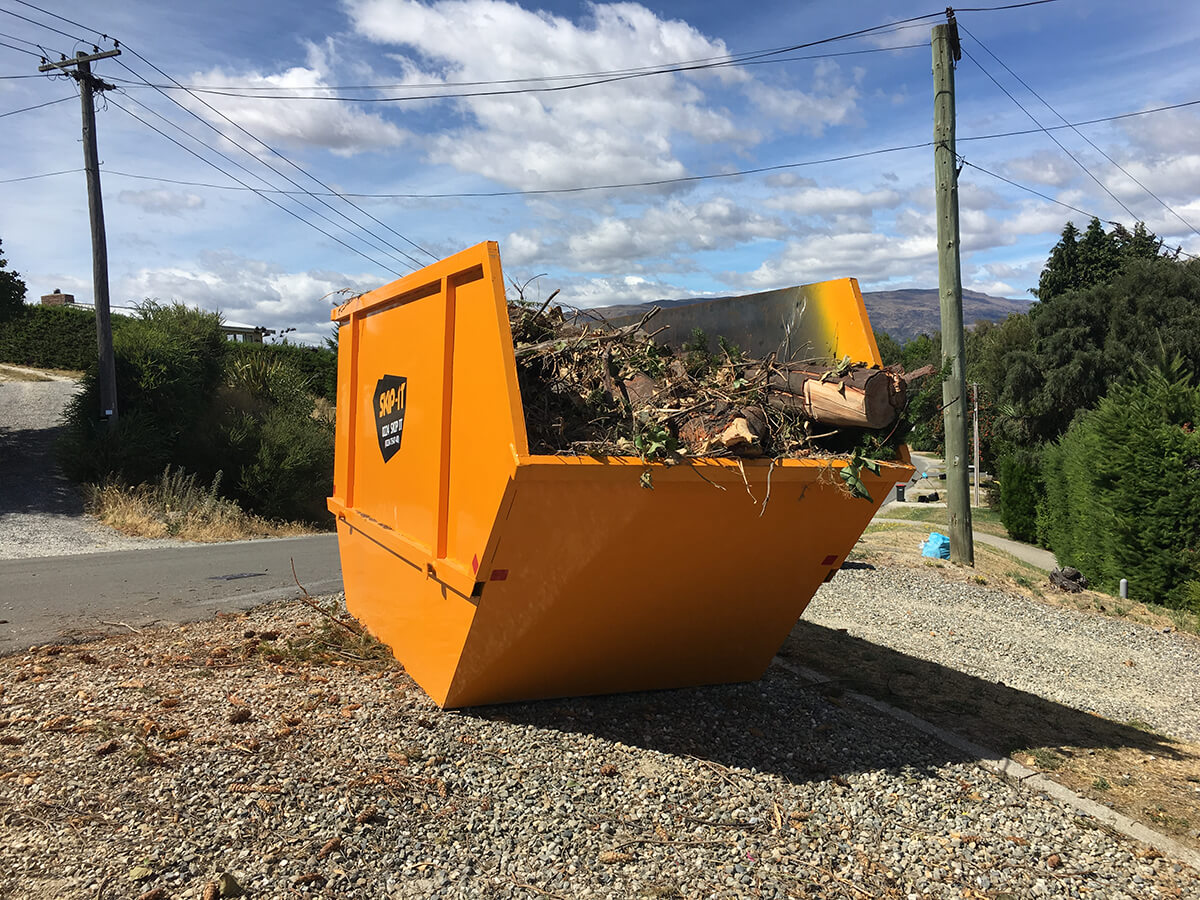 Large orange skip bin filled with branches and tree debris placed on gravel beside a road with power lines overhead and trees in the background under a partly cloudy sky.