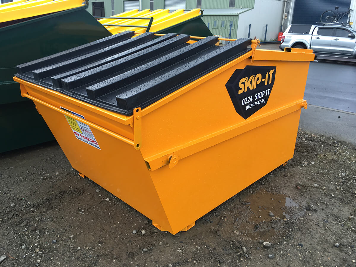 Bright yellow Skip-It brand industrial waste skip with a black ridged lid, placed on gravel outside near green skips and a silver vehicle with a bicycle on its roof rack.
