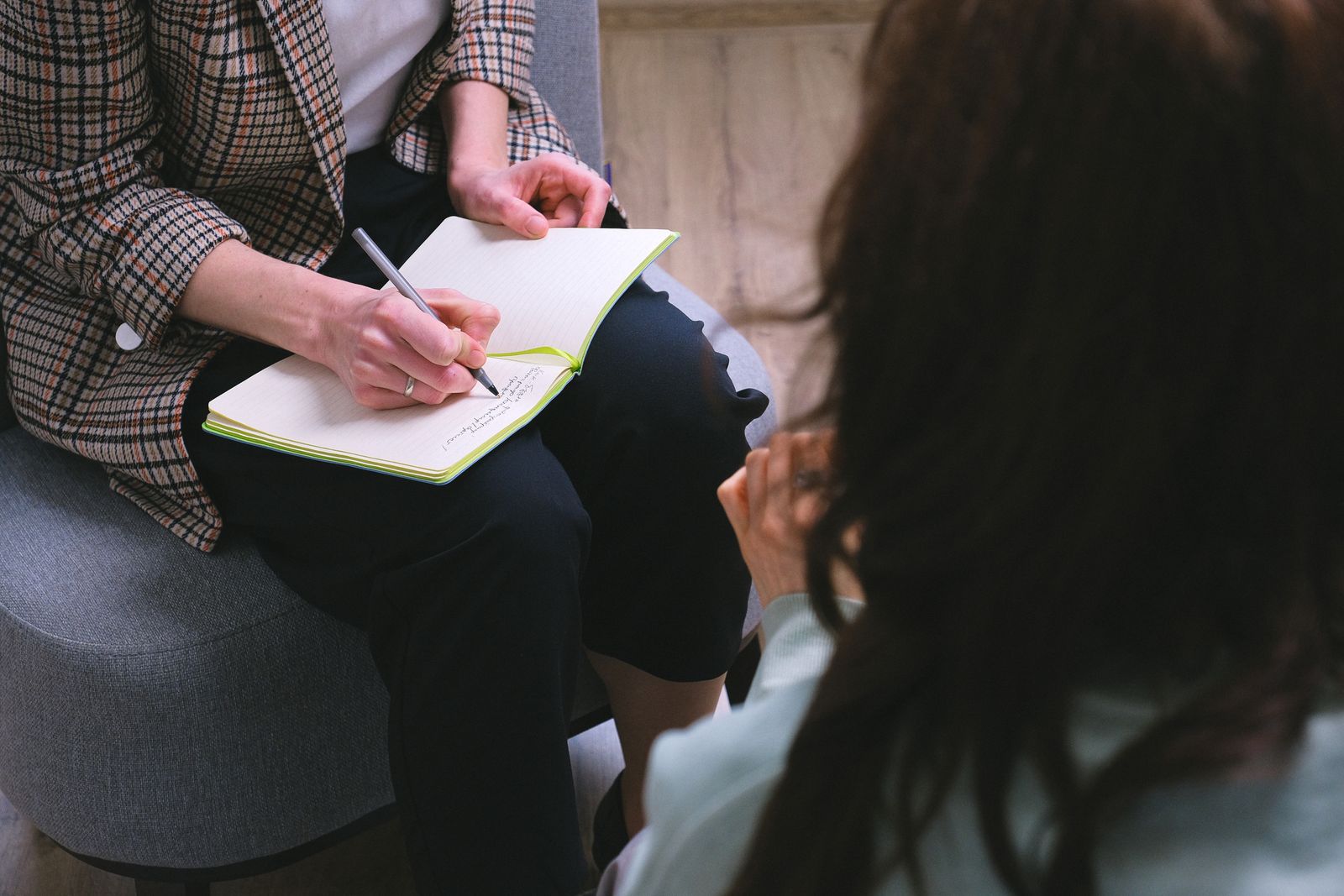 A therapist in a plaid blazer taking notes in a notebook while a client sits across from her