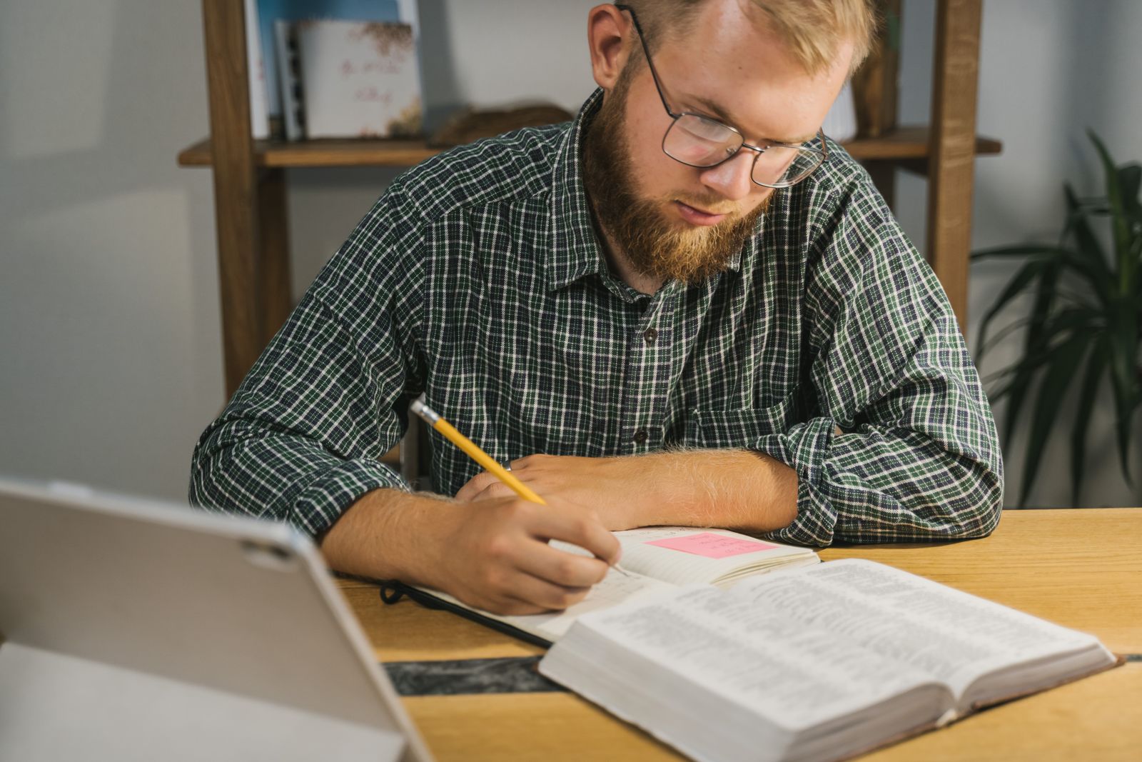 A bearded man with glasses writing in a notebook at a wooden desk beside an open book