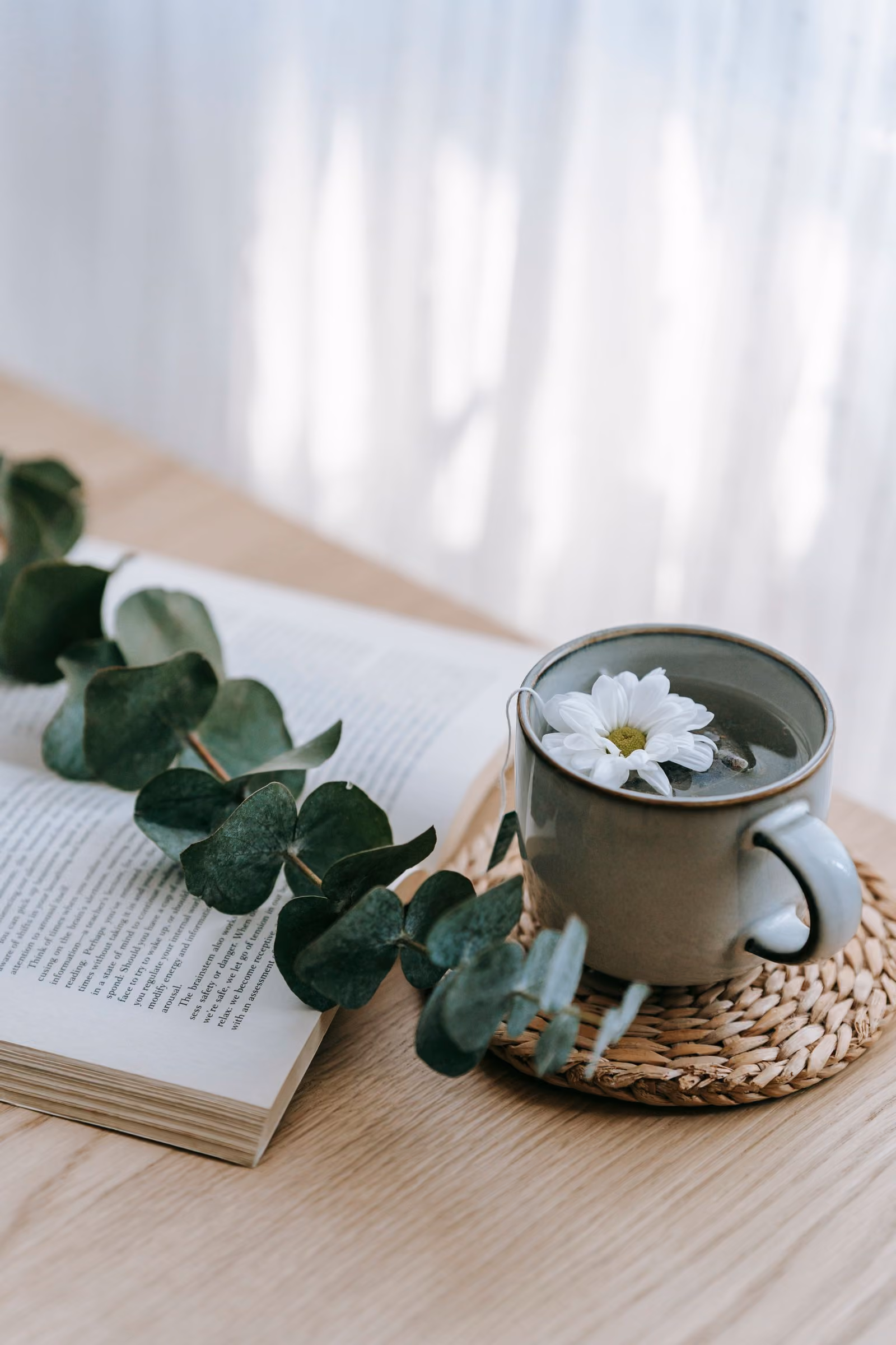 An open book, eucalyptus leaves and a cup of tea with a white flower on a wooden table