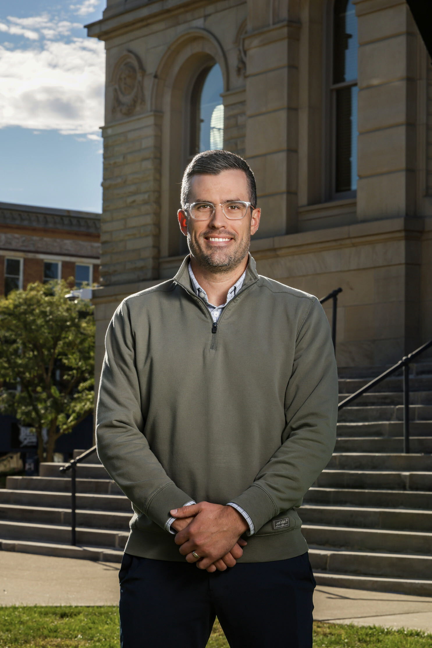 Smiling man with short dark hair and beard, wearing glasses, a green quarter-zip pullover over a checked shirt, standing outdoors in front of stone steps and a building under a partly cloudy sky.
