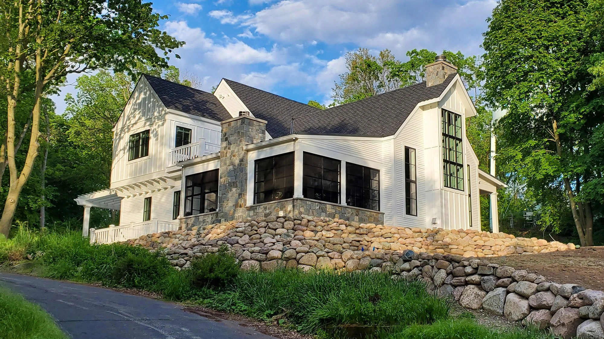 Grand Beach on a Hill residence with white siding, black windows, stone chimneys, and a fieldstone retaining wall