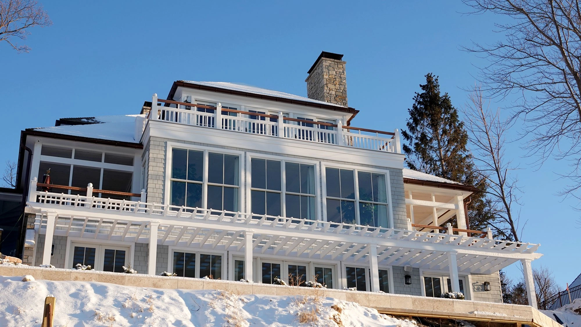 Marquette Compound lake residence with expansive windows, terraces, pergolas, and a stone chimney in winter