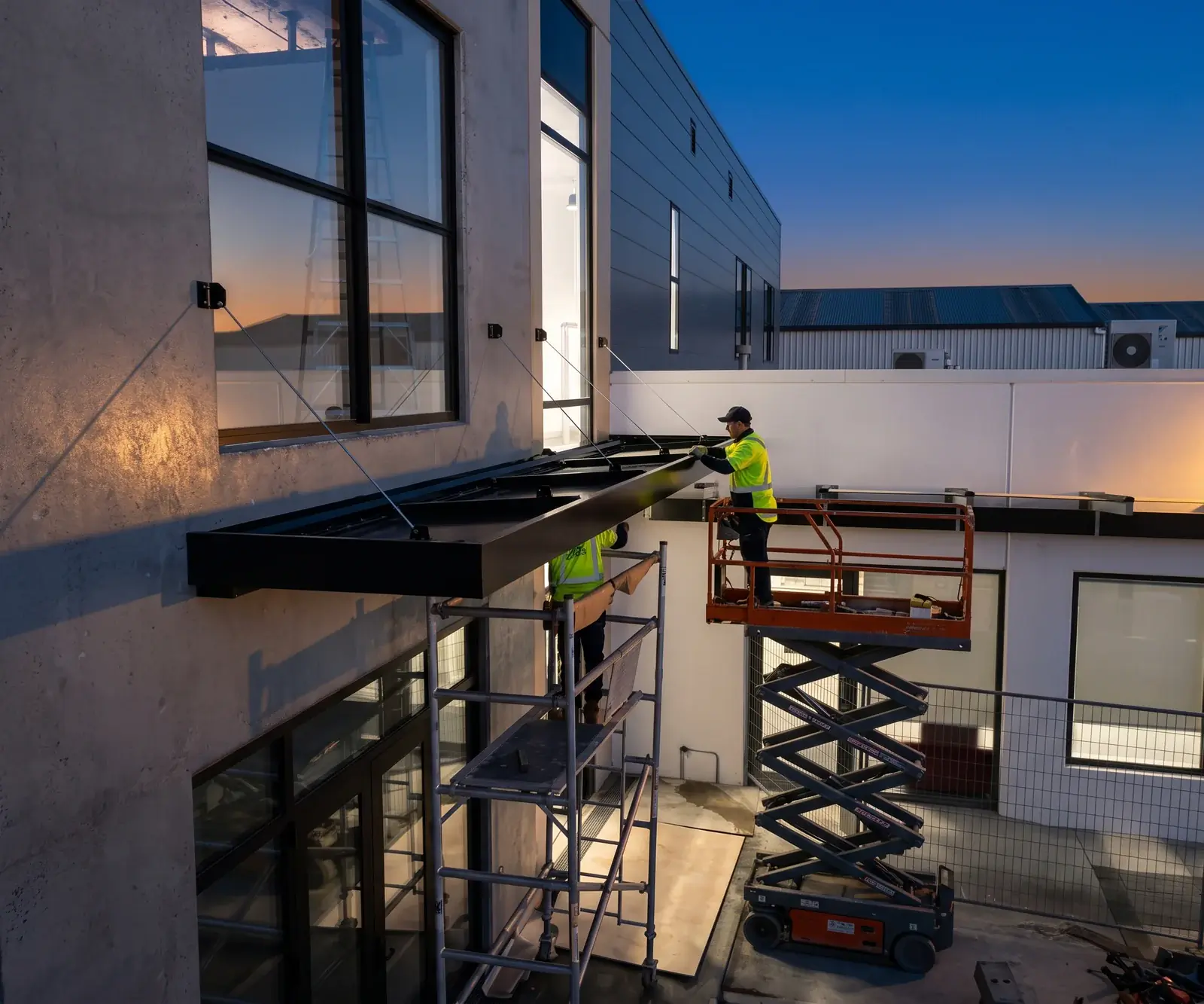Two installers fitting a black entrance canopy from a scaffold and a lift at dusk