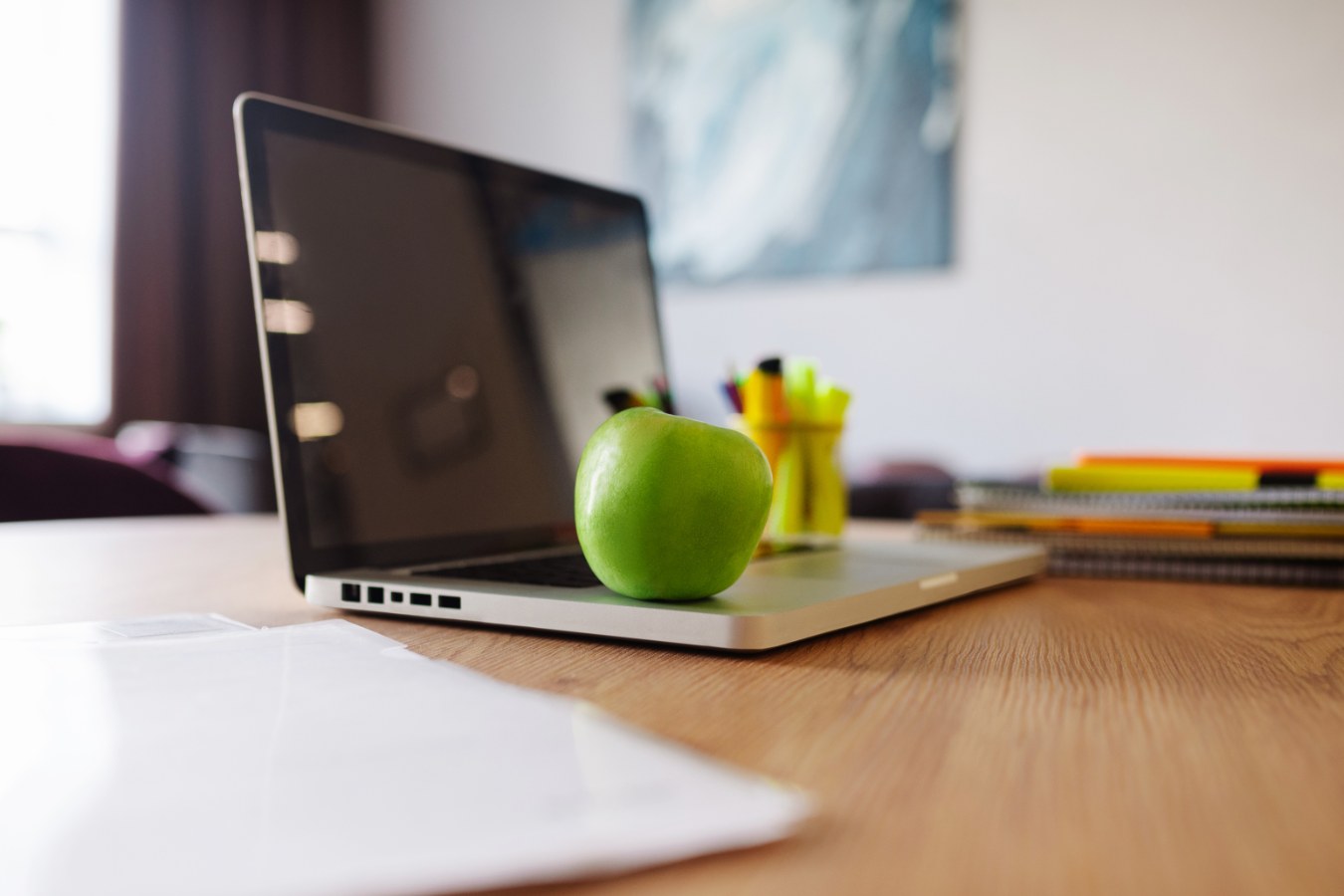 Groene appel ligt op een laptop naast kleurige pennen en schriften