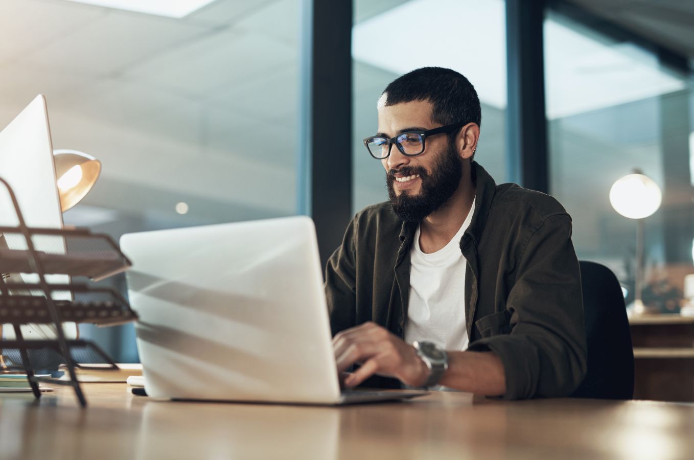Man met bril en baard werkt lachend achter laptop op kantoor