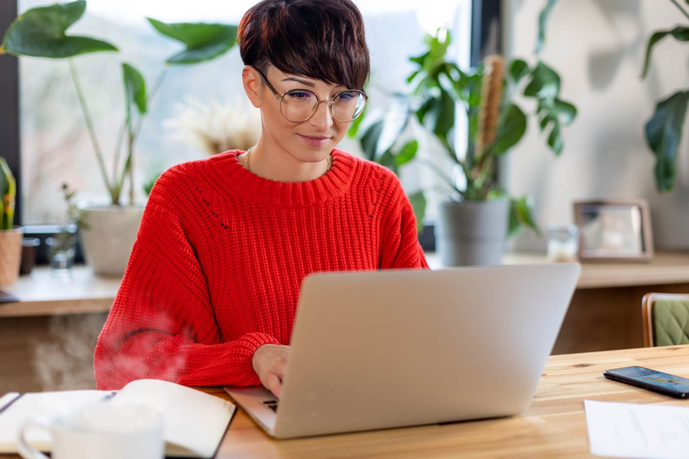 Vrouw met kort haar en rode trui typt op laptop tussen planten