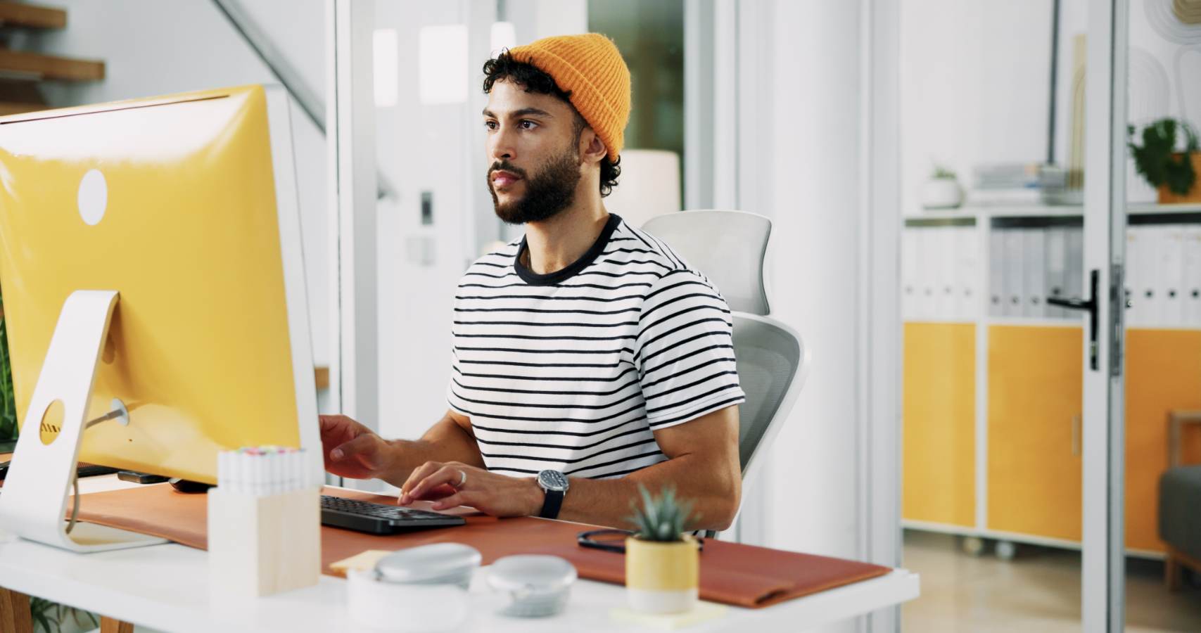 Jonge man met gele muts werkt achter desktopcomputer op kantoor