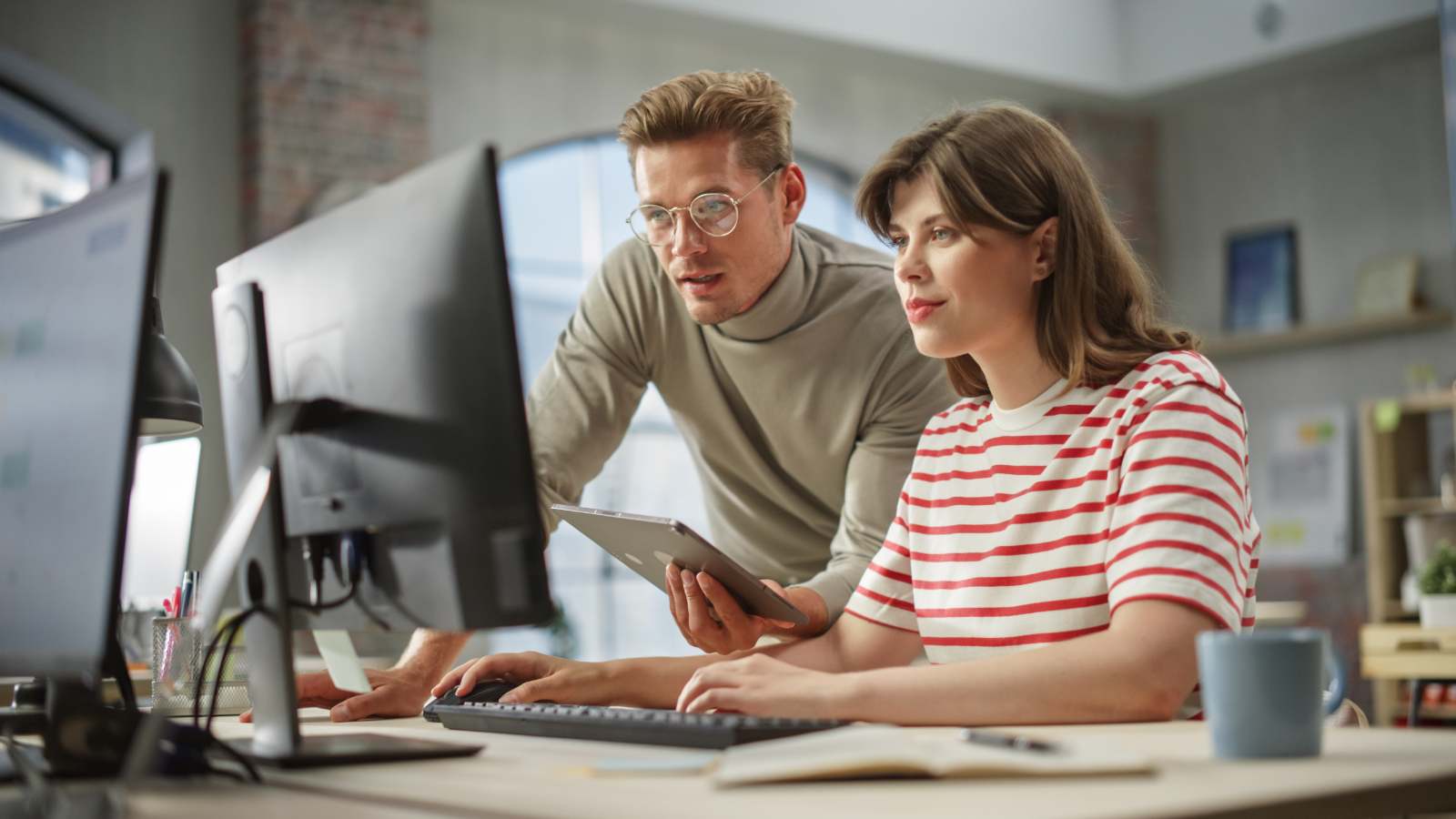 Man instrueert vrouw achter twee computerschermen op kantoor