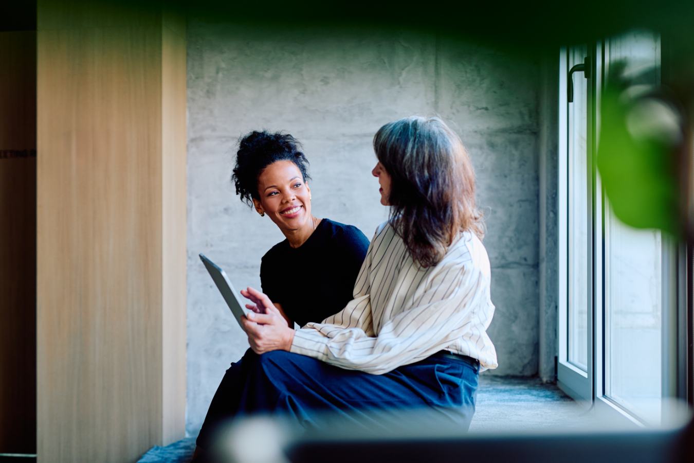 Twee vrouwen bekijken lachend samen een tablet op vensterbank