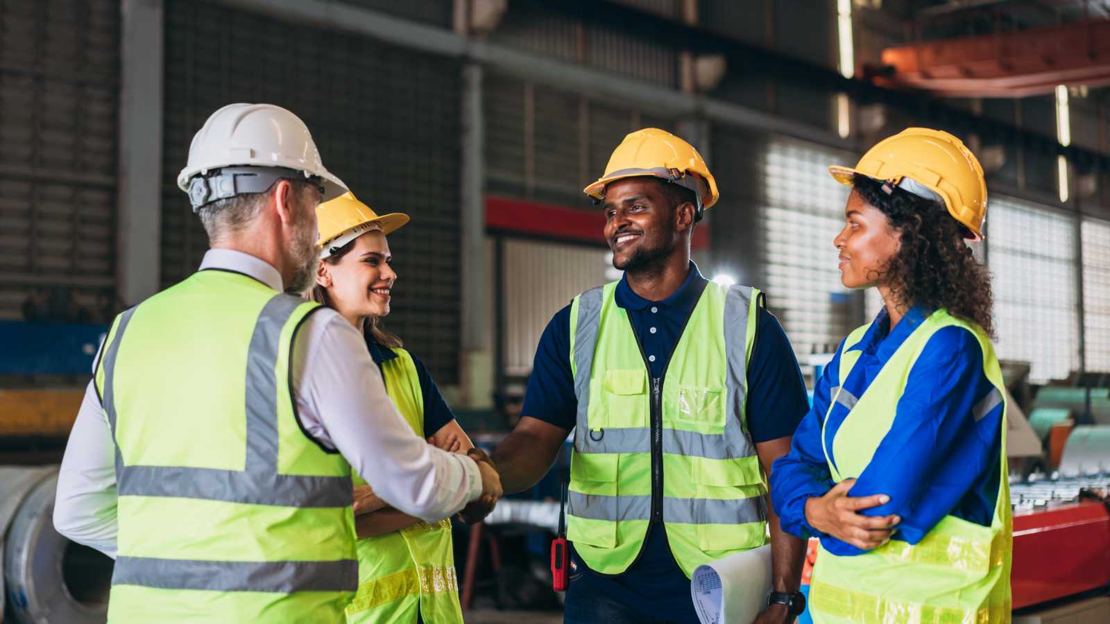 Vier collega's met gele hesjes en helmen begroeten elkaar in fabriek