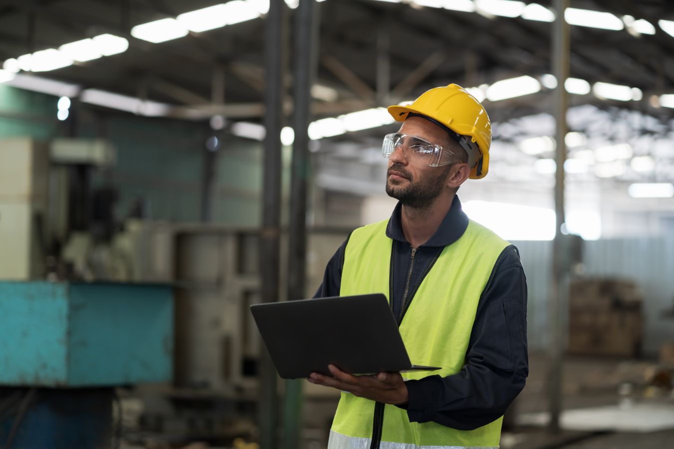 Monteur met gele helm en veiligheidsbril werkt met laptop in fabriekshal