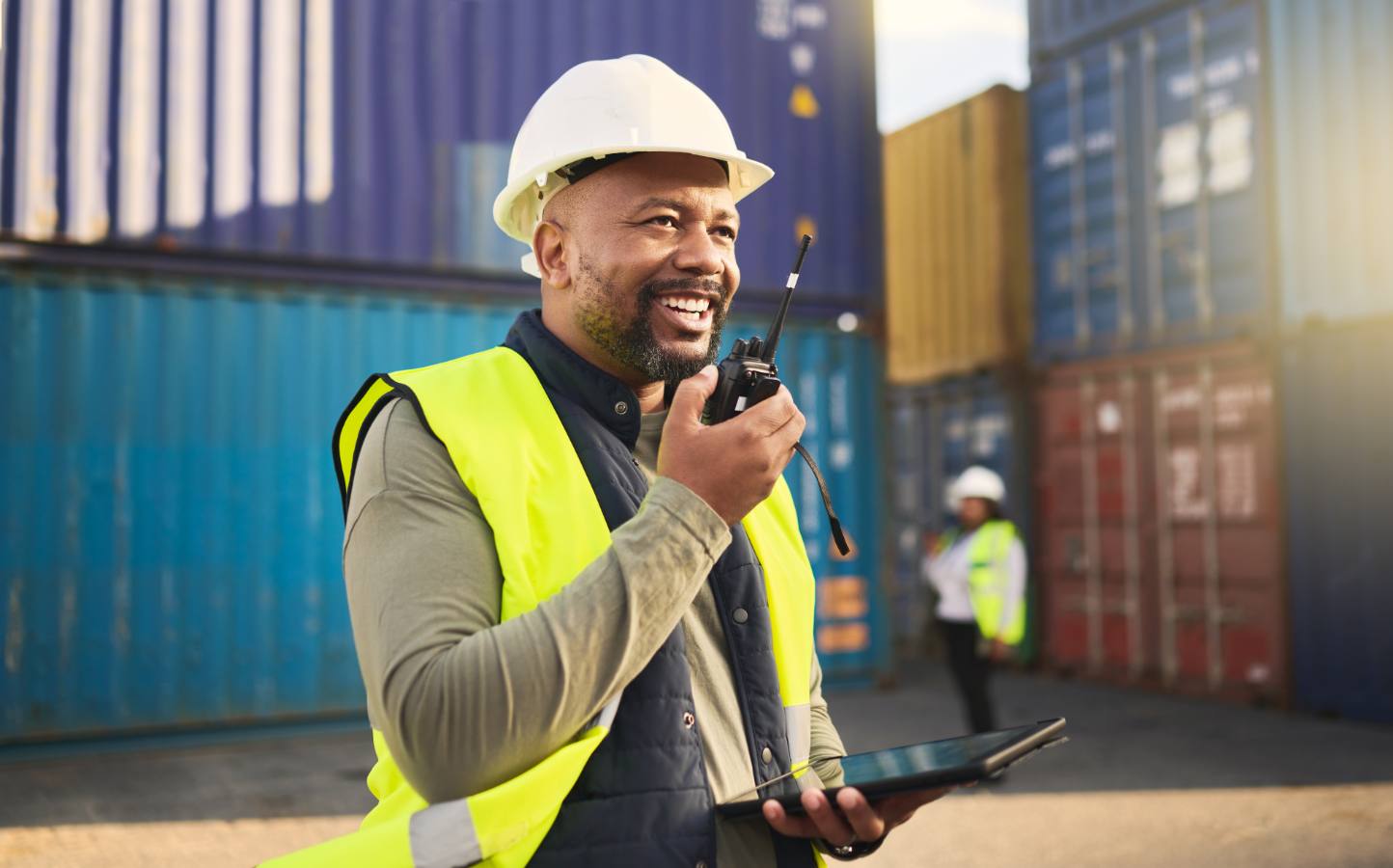 Man met helm en veiligheidsvest praat via een portofoon bij containers