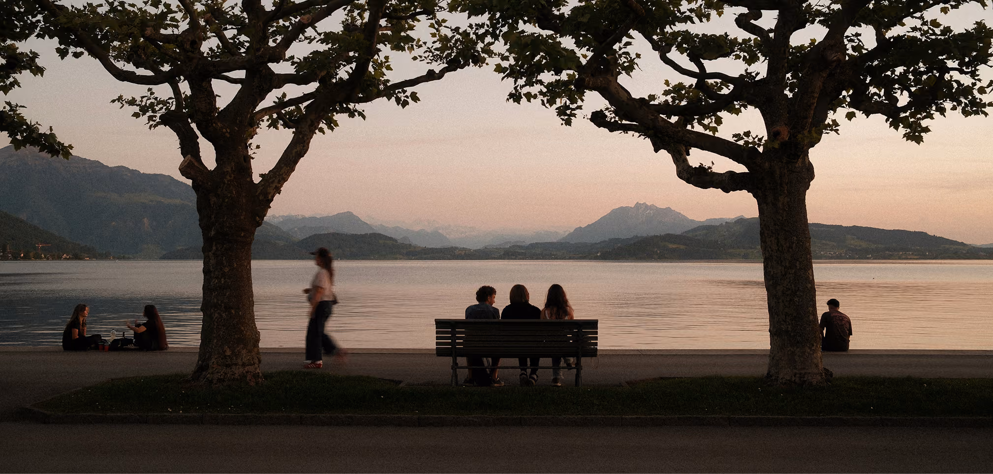 Three people sitting on a bench facing a calm lake with mountains in the distance at sunset, framed by two leafy trees, while others sit or walk nearby on the lakeside promenade.