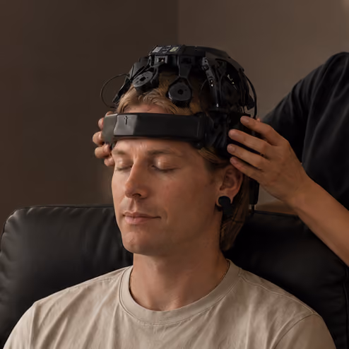 Man sitting with eyes closed wearing a black EEG headset while another person adjusts it on his head.