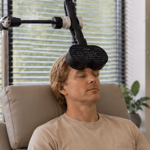 Blond man with closed eyes sitting in a beige recliner while a black device with multiple sensors is positioned on his forehead, attached to a mechanical arm in a room with window blinds and a green plant in the background.