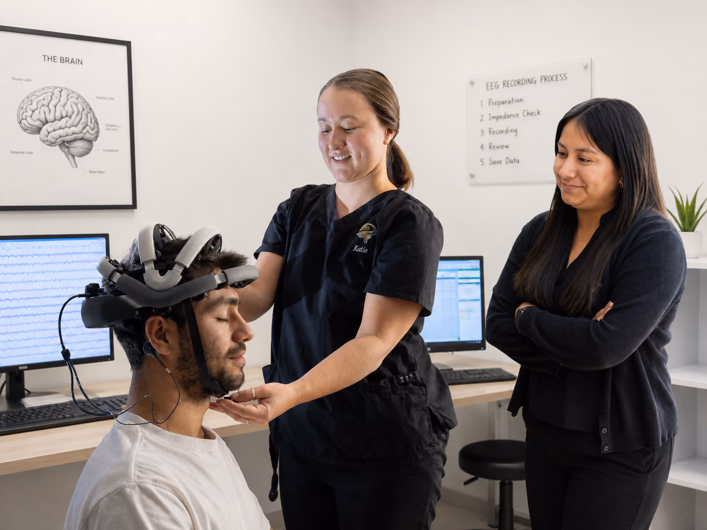 A medical professional in black scrubs adjusts a brain-monitoring device on a seated man's head while another woman watches in a clinical office with brain diagrams and EEG recording process steps on the walls and computer monitors displaying EEG data.