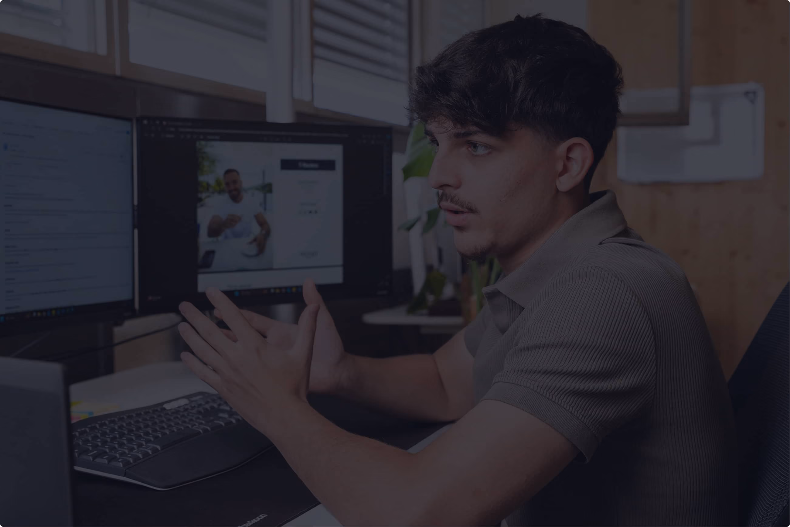 Junger Mann in einem grauen Poloshirt sitzt an einem Schreibtisch mit zwei Computermonitoren und spricht gestikulierend, während er in einem modernen Büro arbeitet.